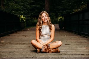 A smiling young woman sits cross-legged on a wooden bridge wearing a plaid dress and brown boots.