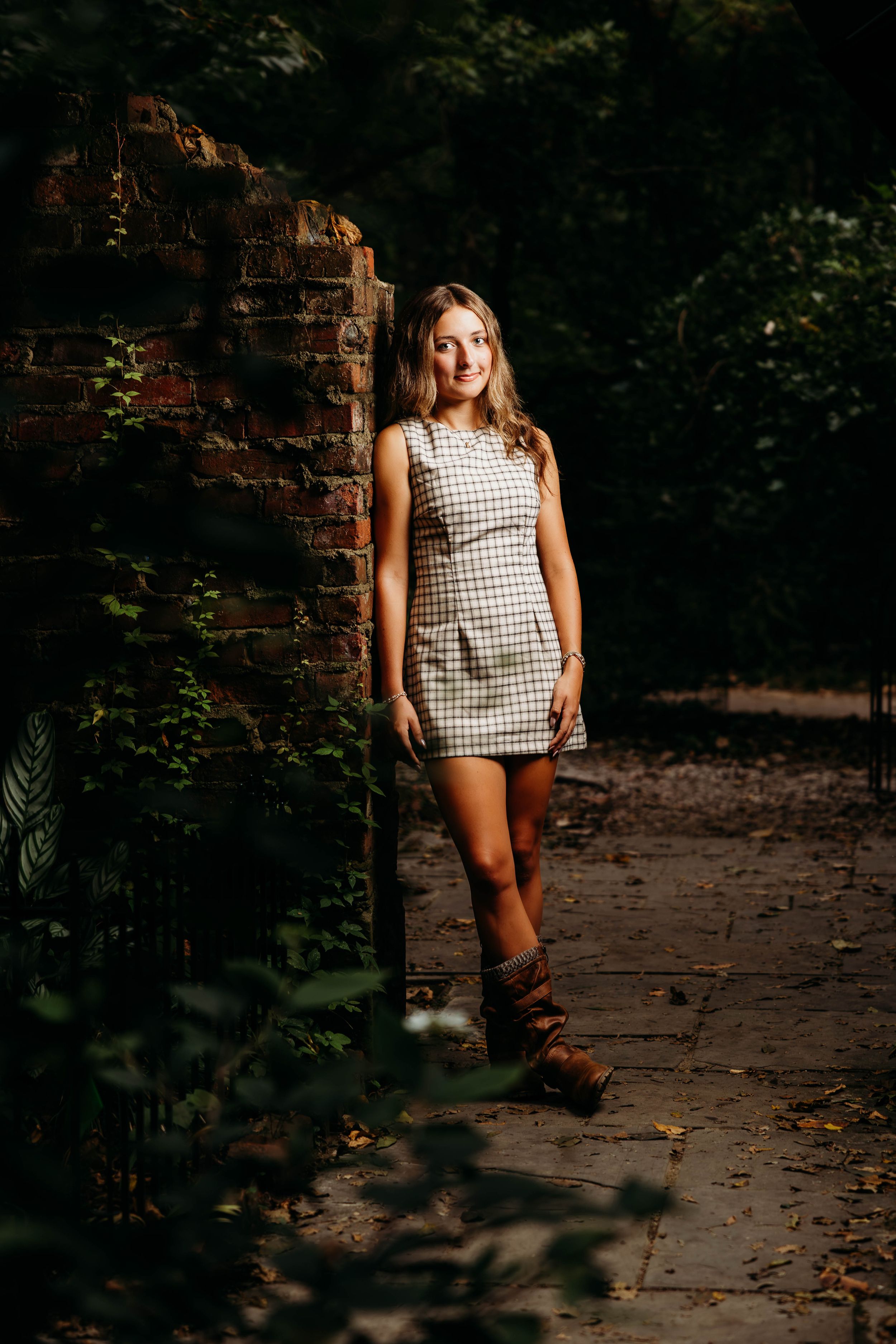 Young woman in plaid mini dress and brown boots leaning against brick wall in lush garden pathway.