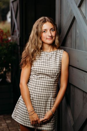 Young woman in plaid mini dress leaning against rustic gray barn door, long wavy hair, gold bracelets.