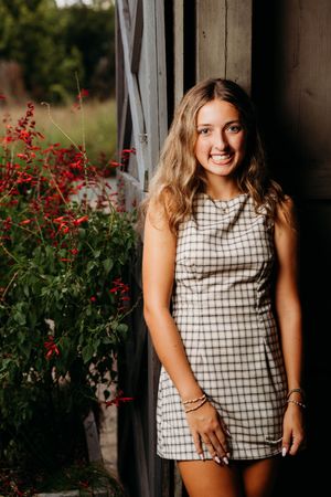 Young woman in plaid mini dress smiling outdoors near red flowers and rustic wooden wall.