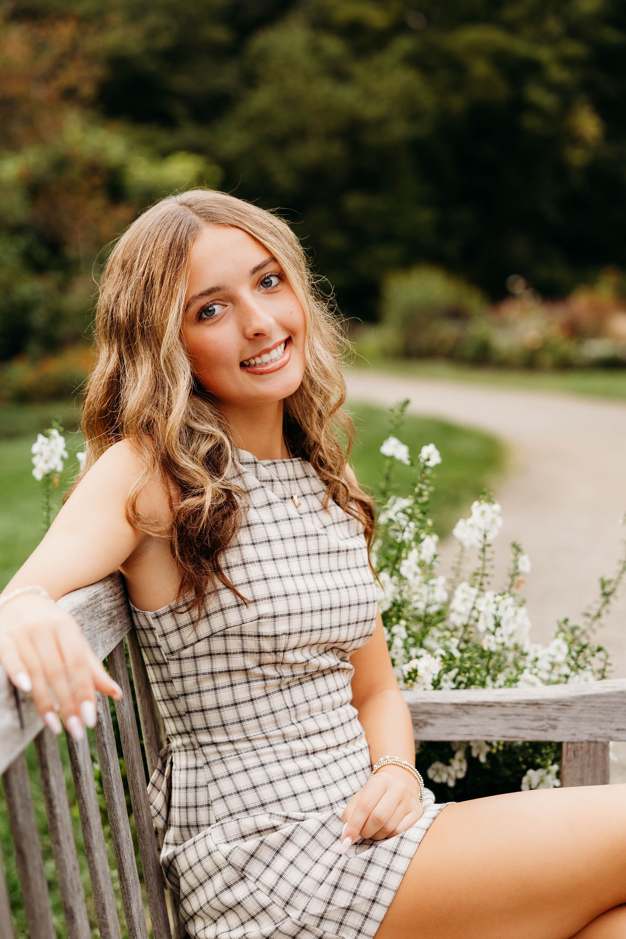 Young woman in plaid dress smiling on wooden bench surrounded by white flowers in a lush garden setting.
