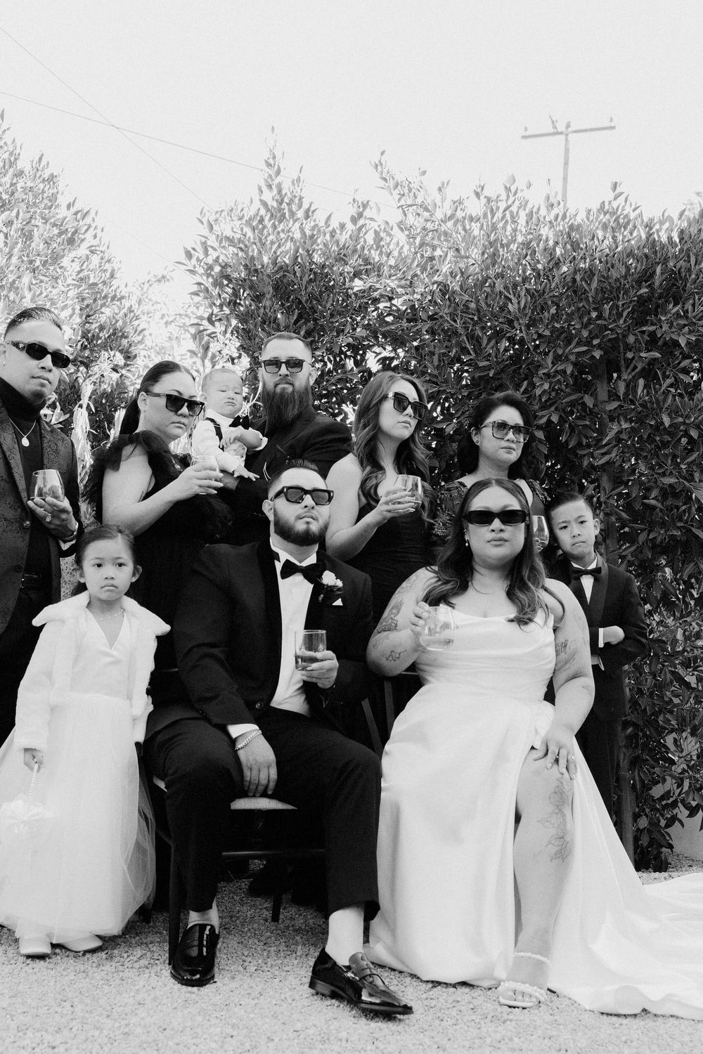 Black and white wedding photo of bride and groom with wedding party wearing sunglasses, posing outdoors near hedges.