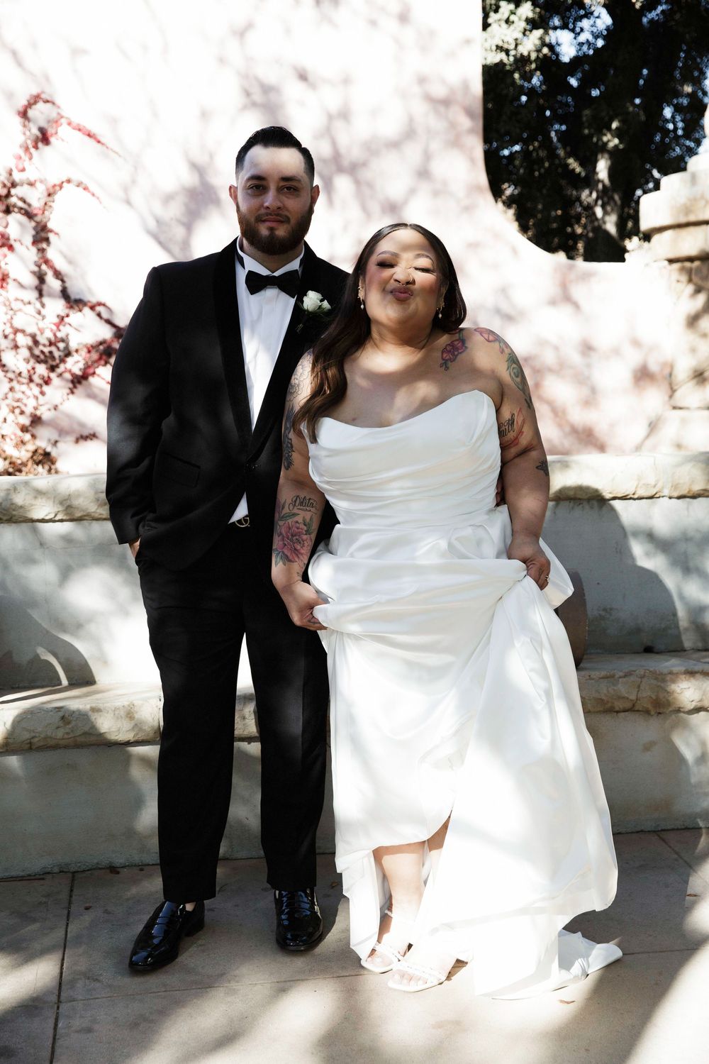 A bride in a white strapless gown and groom in a black tuxedo pose together outdoors on their wedding day.
