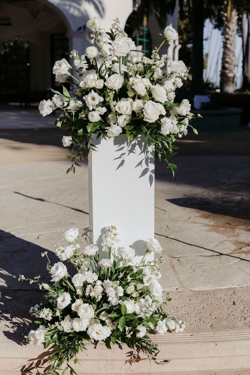 White floral arrangement with roses and greenery on a white pedestal for an elegant outdoor wedding ceremony.