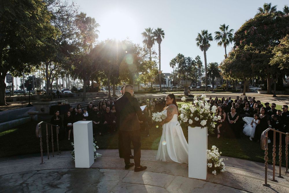 Outdoor wedding ceremony at sunset with couple at altar, white floral arrangements, palm trees, and seated guests in background.