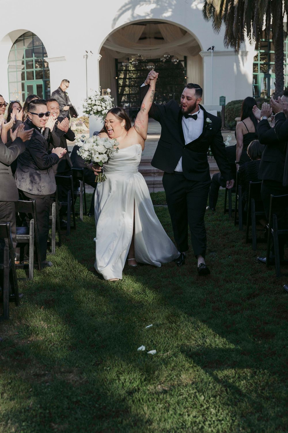 Joyful newlywed couple raises hands walking down outdoor aisle after wedding ceremony, bride holding white bouquet.