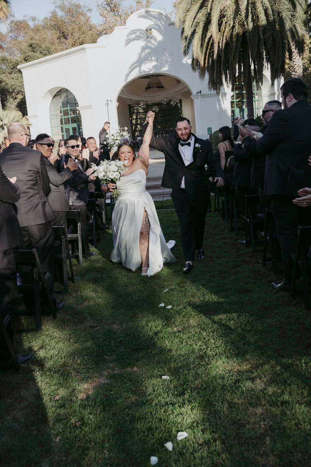 Bride and groom celebrate joyfully walking down outdoor aisle after wedding ceremony, guests applauding beside white chapel.