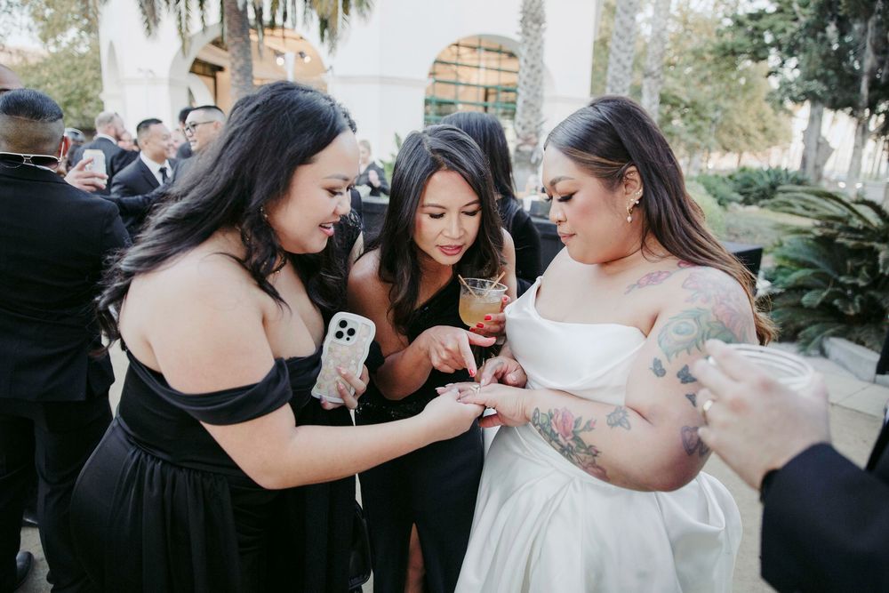 Bride in white gown with tattooed arms shares a moment with bridesmaids in black dresses at outdoor wedding reception.