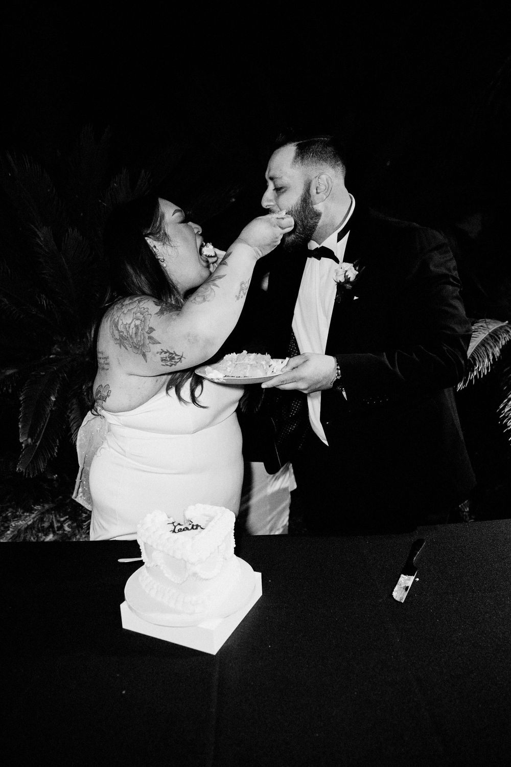 Black and white photo of bride feeding wedding cake to groom at reception, with tiered cake on table.