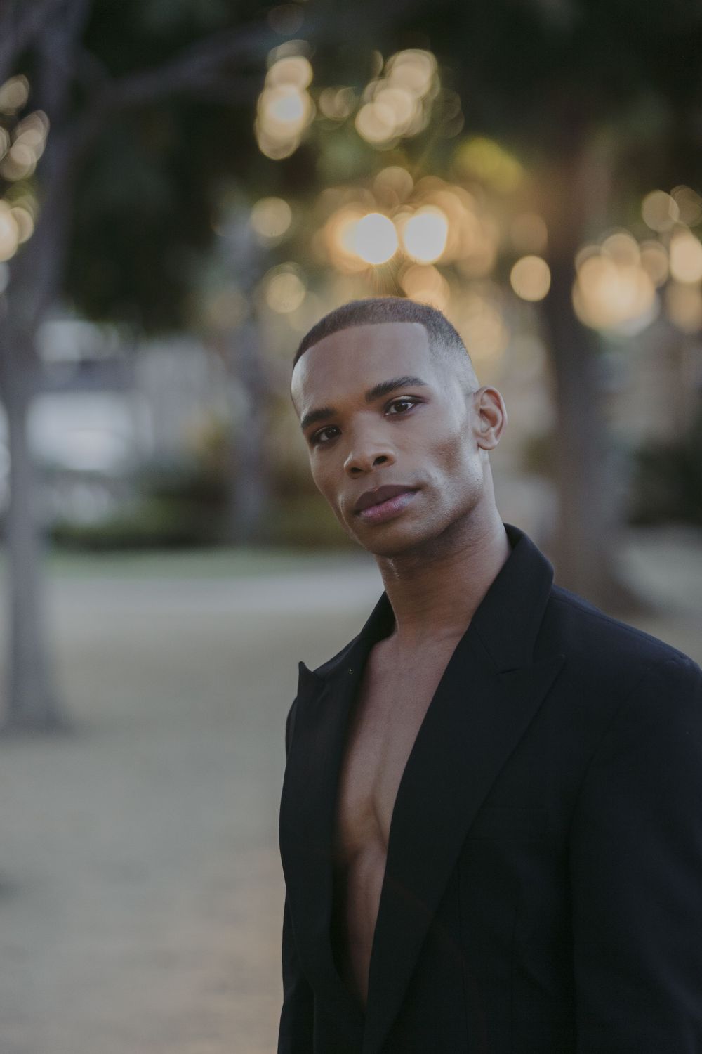 Young Black man in open black blazer poses outdoors at dusk with bokeh lights in background.