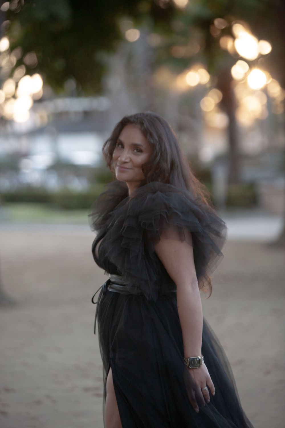 Woman in elegant black tulle off-shoulder gown posing outdoors at dusk with bokeh lights in background.