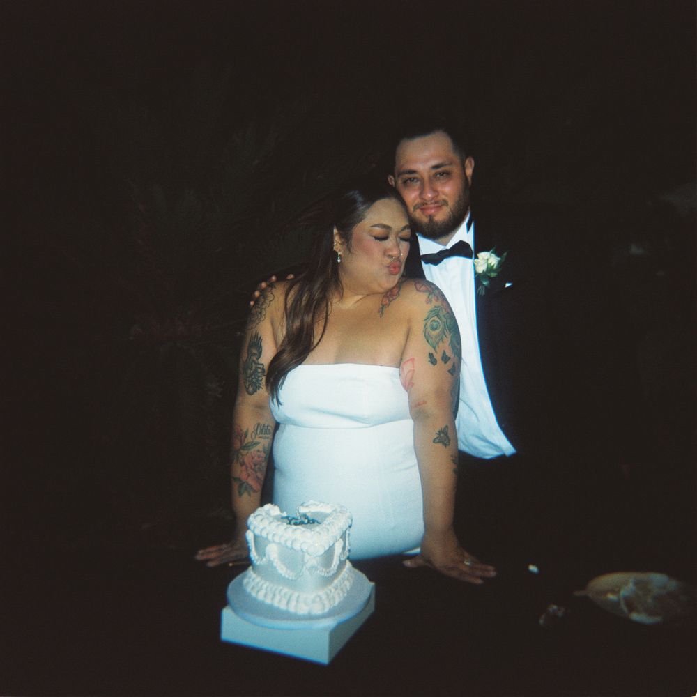 Bride and groom pose together behind a white wedding cake at their nighttime outdoor wedding reception.