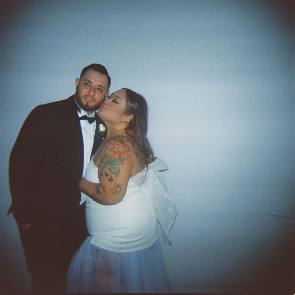 A couple in wedding attire poses romantically, groom in tuxedo and bride in white dress with floral shoulder tattoo.