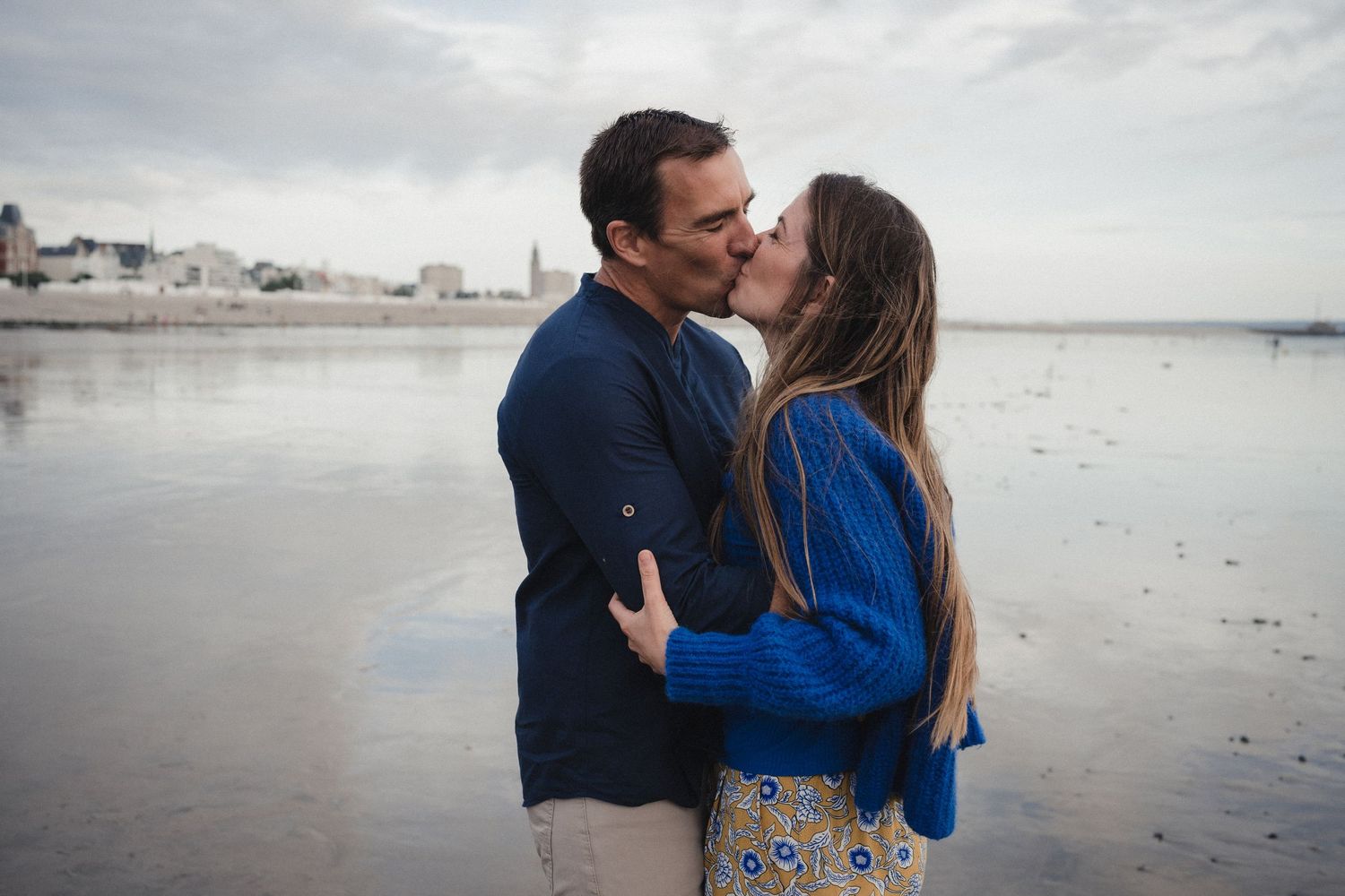 Couple sharing a romantic kiss on a beach with a coastal town skyline in the background on a cloudy day.