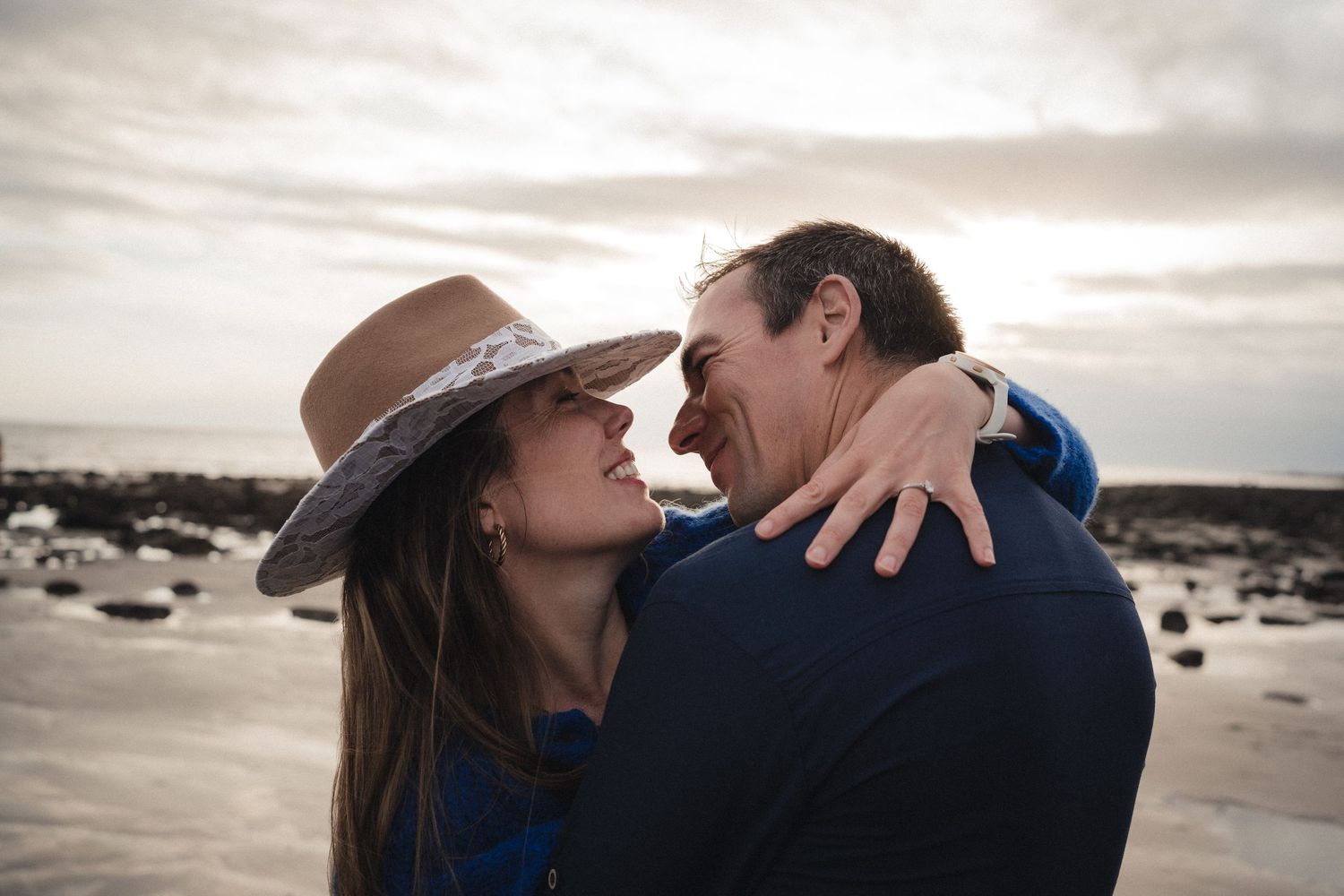 Romantic couple sharing a tender moment on a beach, woman in wide-brim hat embracing man under cloudy sky.