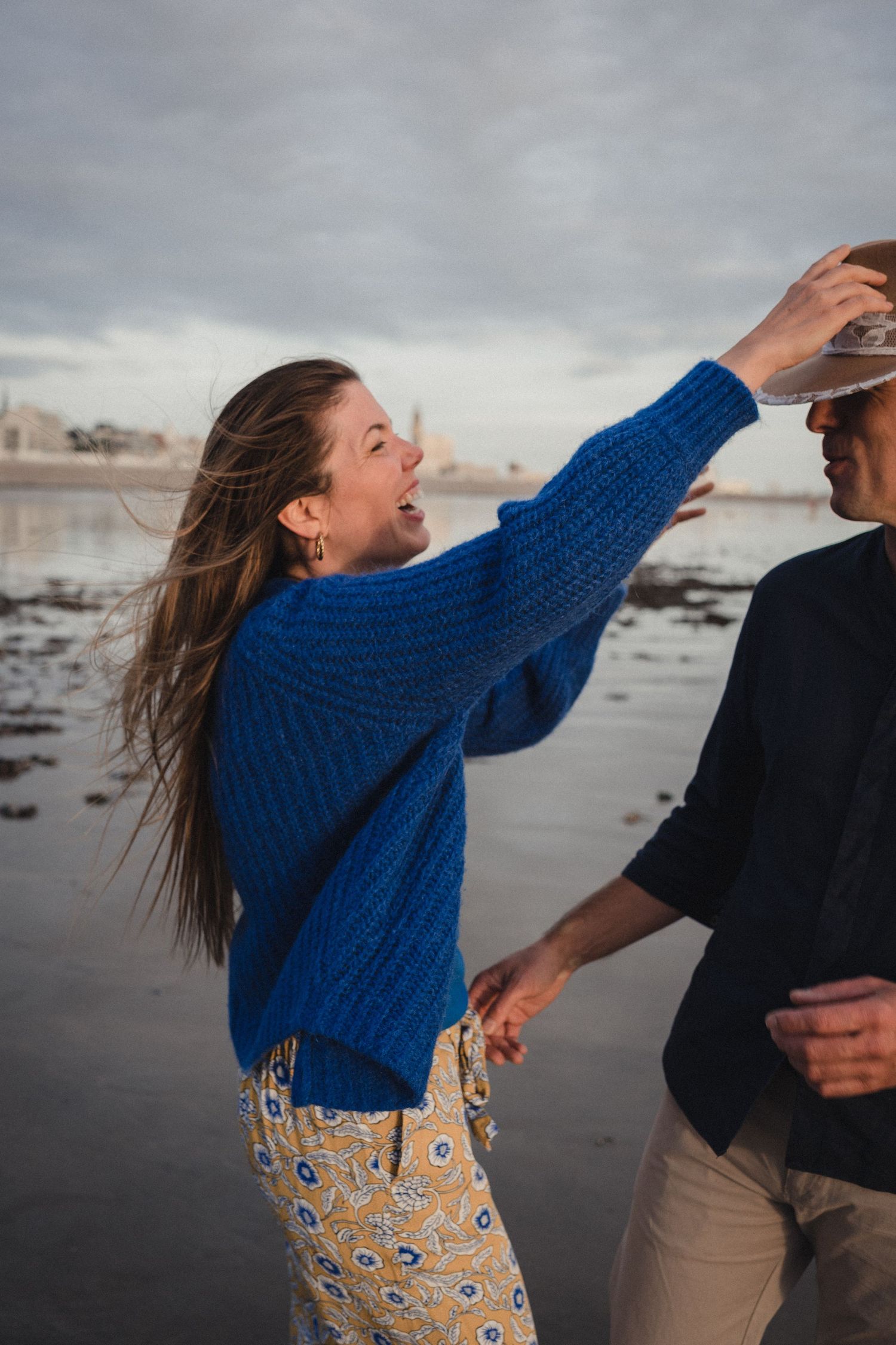 A woman in a blue knit sweater and floral pants playfully adjusts a man's hat on a cloudy beach.