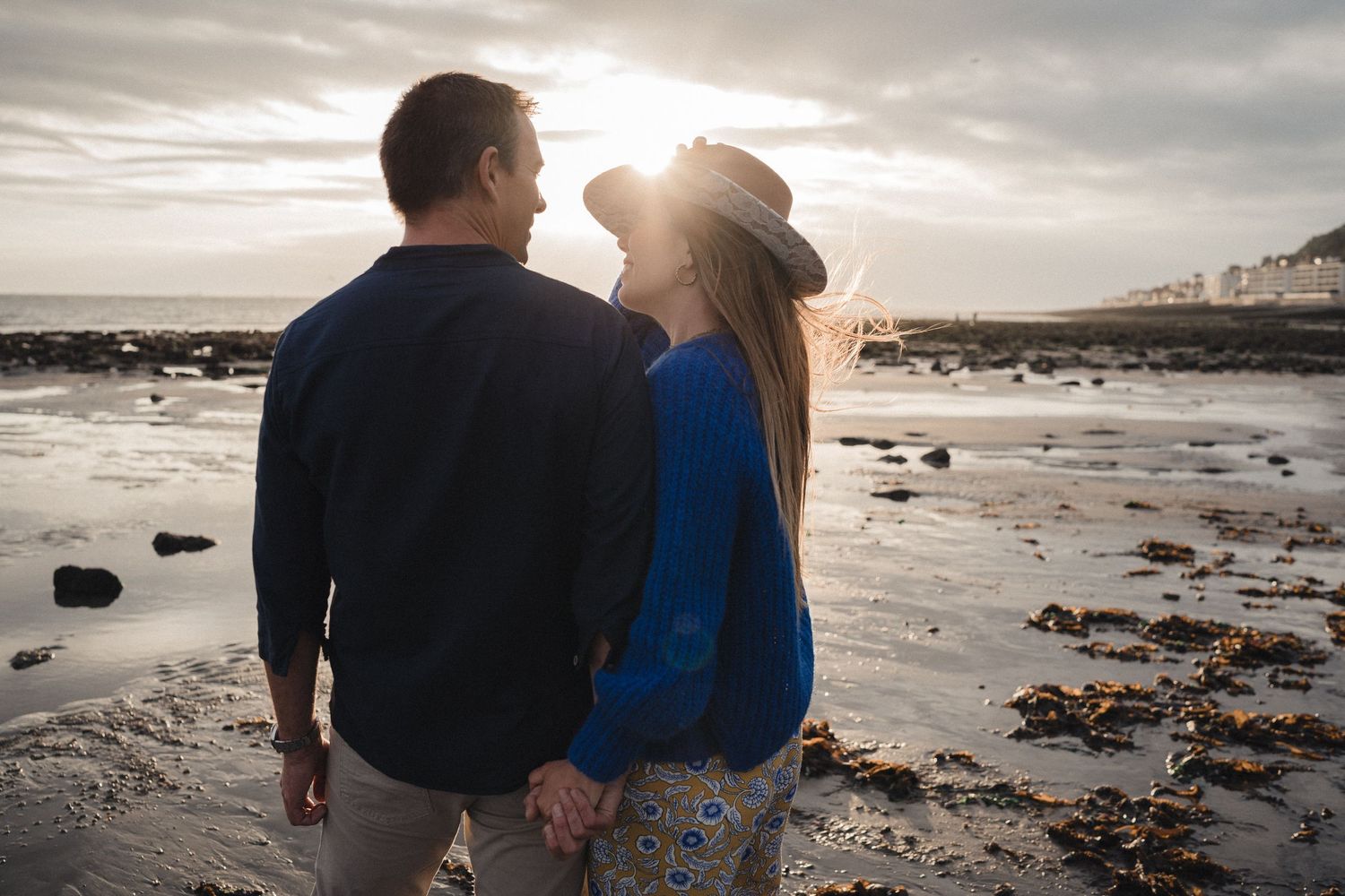 Couple holding hands on a rocky beach at sunset, woman wearing blue sweater and hat.