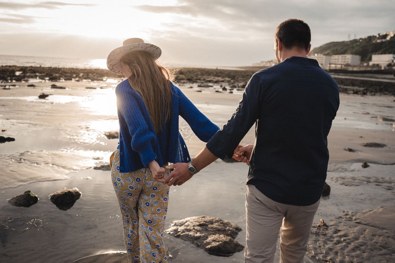 Couple holding hands walking on a scenic beach at sunset, woman in blue jacket and floral pants wearing a hat.