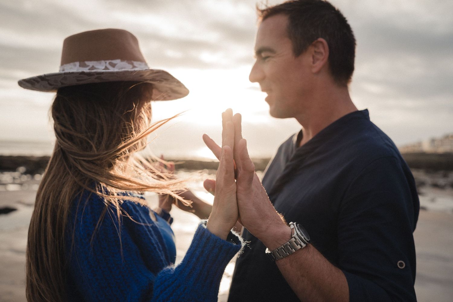 Couple sharing a tender moment at sunset on a beach, woman in blue sweater and hat facing man in navy shirt.