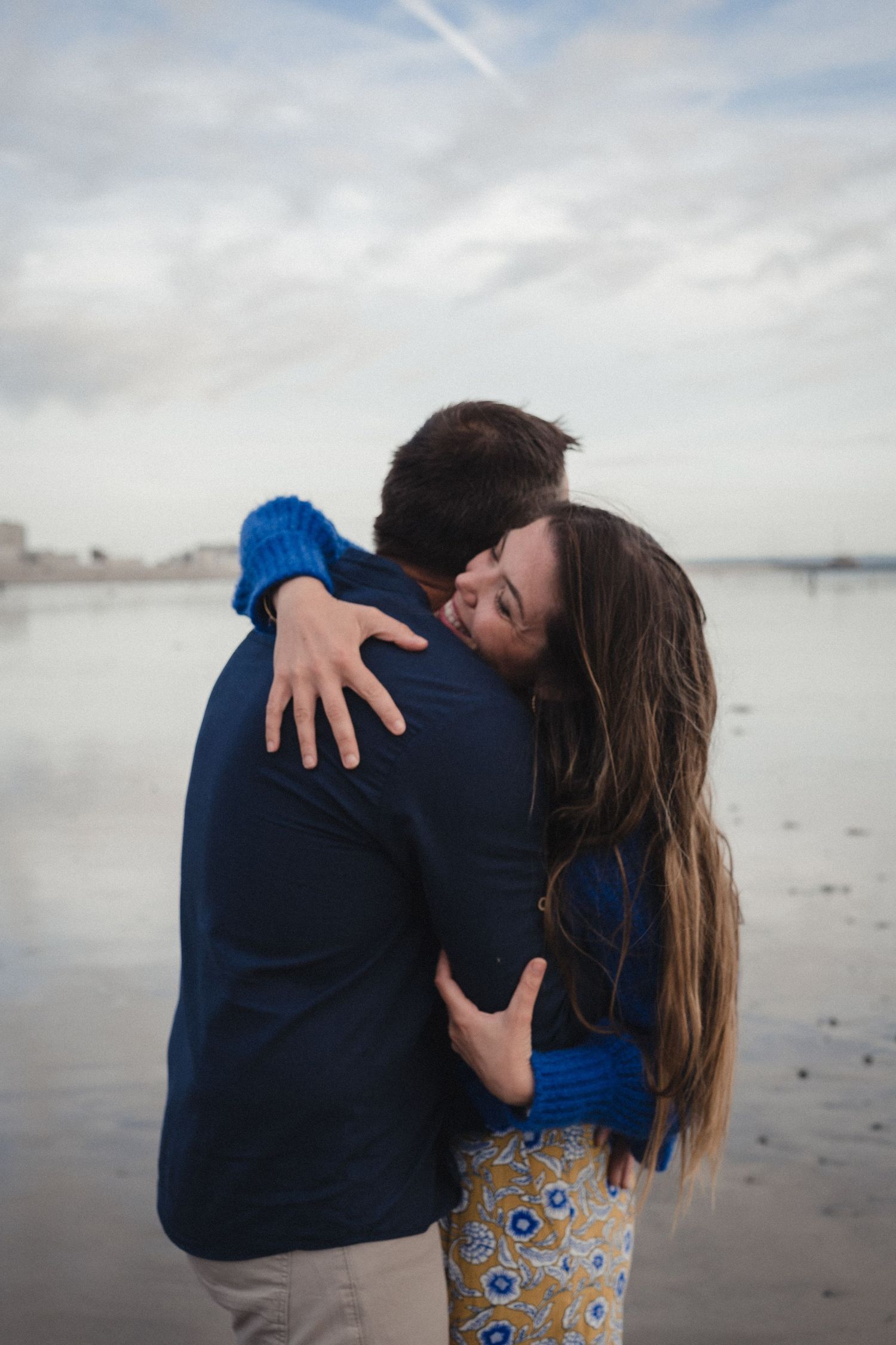 Couple embracing on a beach, woman in blue sweater and floral pants hugging man in navy jacket by calm water.