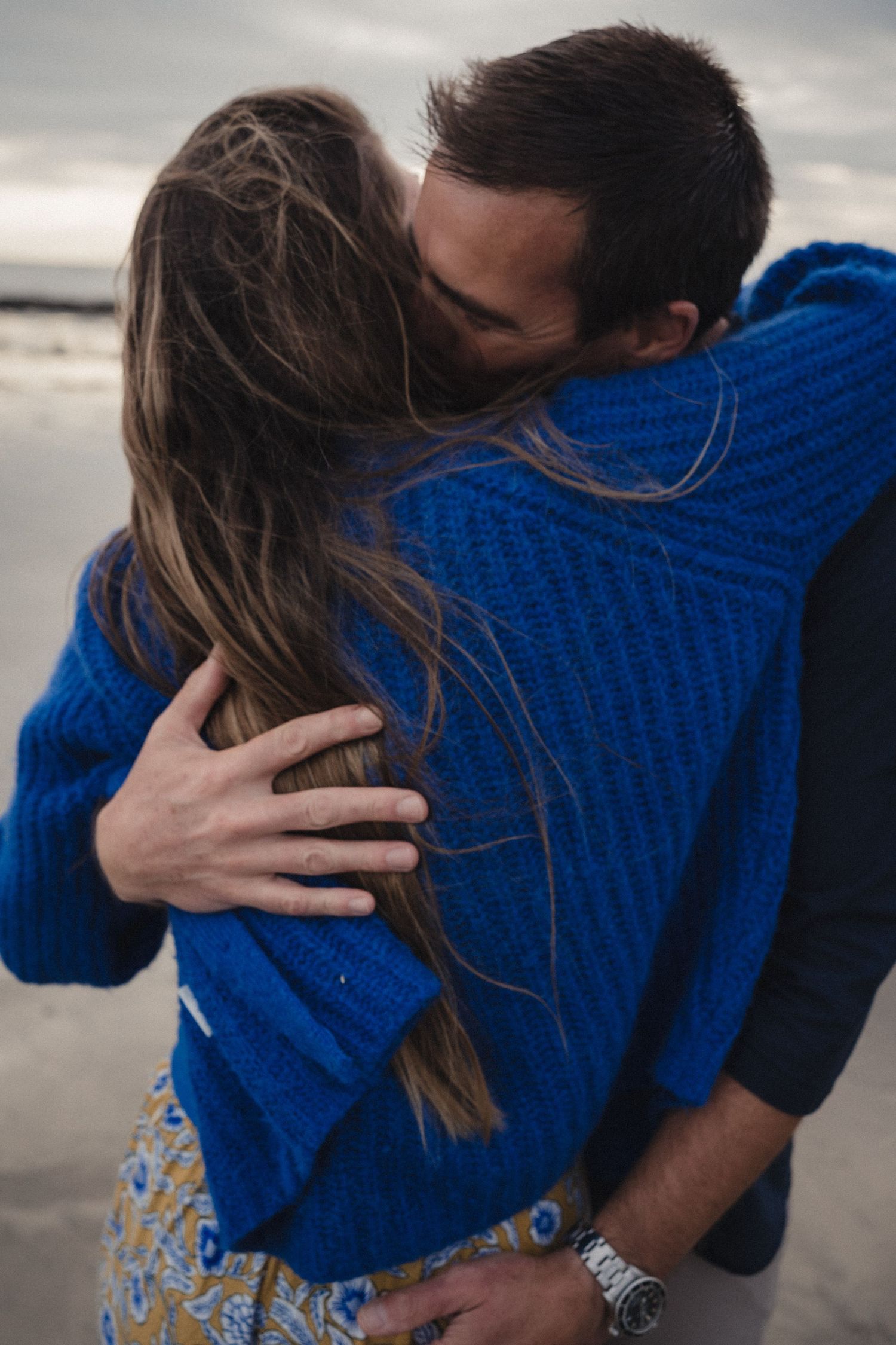 Couple embracing on a beach, wearing blue knit sweaters, sharing a tender moment with ocean in the background.