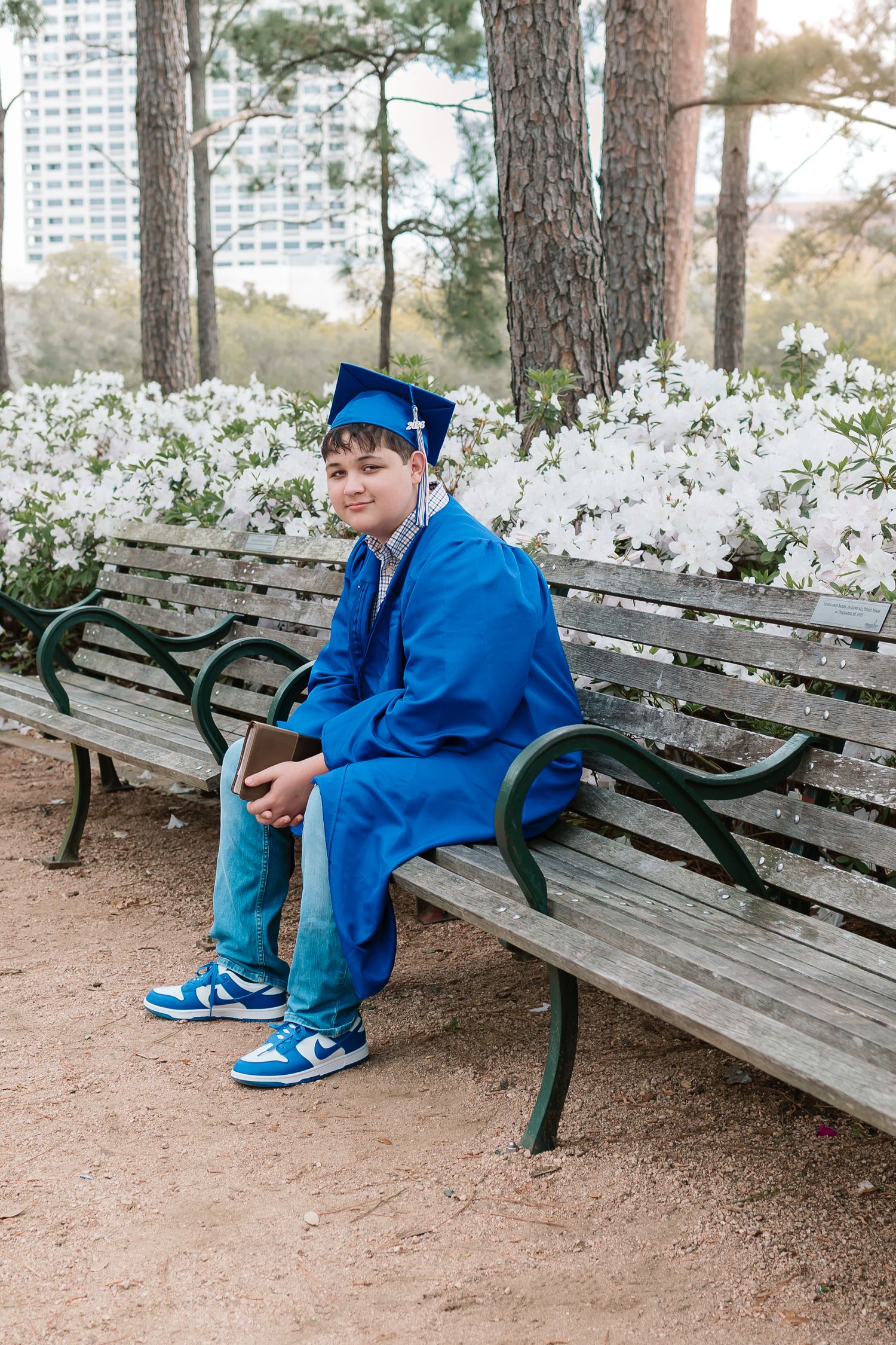 Graduate in blue cap and gown sits smiling on a park bench surrounded by white flowering bushes.