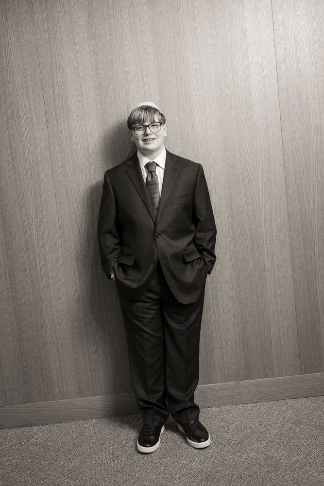 Young man in dark suit, tie, glasses, and yarmulke stands confidently against wood-paneled wall in black and white photo.