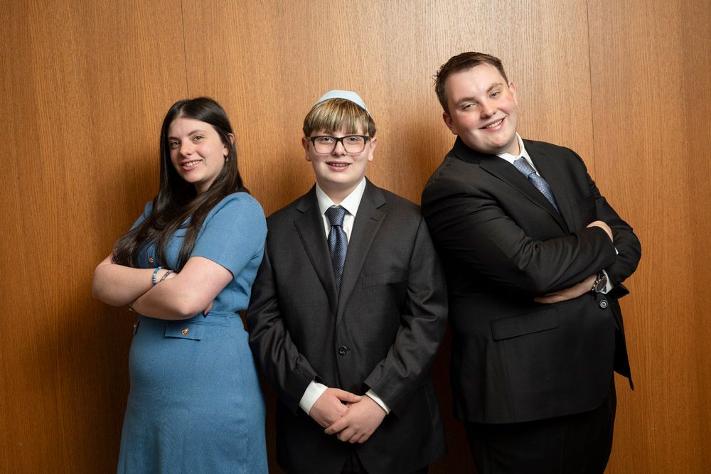 Three formally dressed young people posing together, smiling, against a wooden wall backdrop.