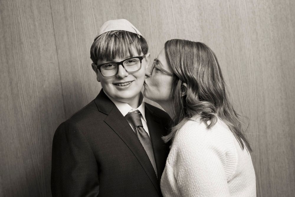 Black and white photo of a young man in a kippah and suit receiving a kiss on the cheek from a woman.