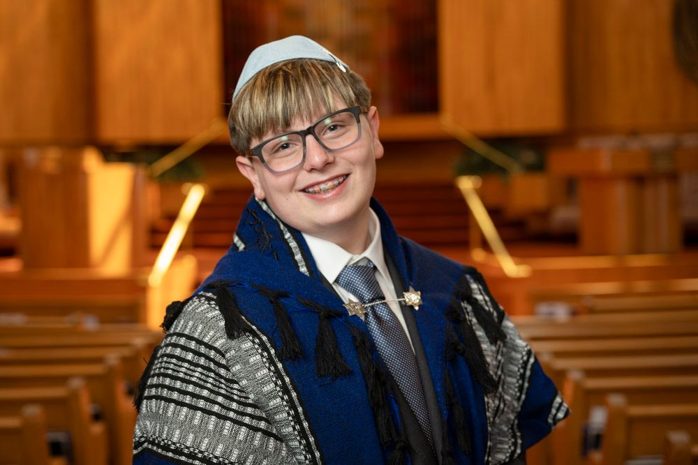 Young boy in tallit and kippah smiling during Bar Mitzvah ceremony inside a synagogue.