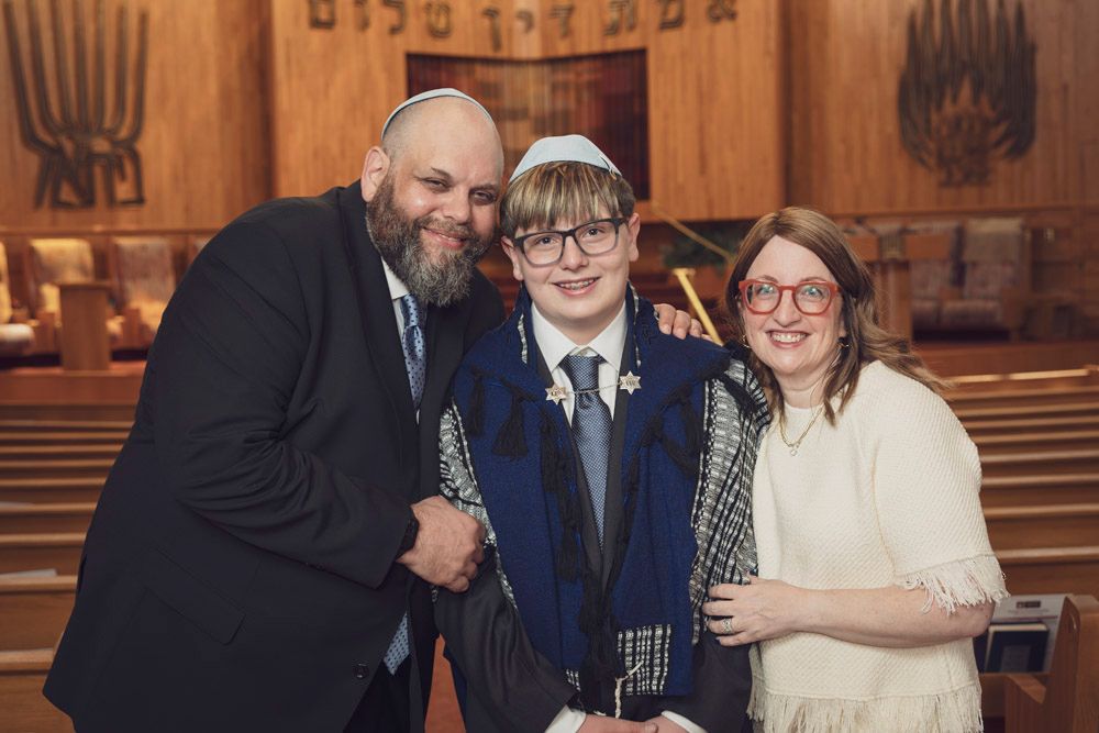 Family celebrating a Bar Mitzvah in a synagogue, boy wearing tallit and kippah flanked by proud parents.