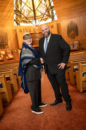Two people pose together in a synagogue during a Bar Mitzvah ceremony, one wearing a tallit prayer shawl.