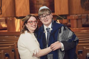 A boy in a kippah and tallit smiles with a woman in red glasses during a Bar Mitzvah ceremony in a synagogue.