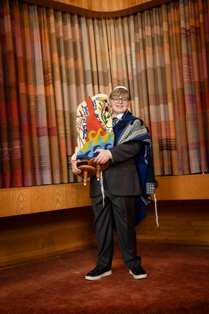 Young man in suit and tallit holds Torah scroll during Bar Mitzvah ceremony in synagogue.