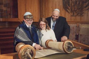Rabbi guides two children reading from Torah scroll during Bar Mitzvah ceremony in synagogue.