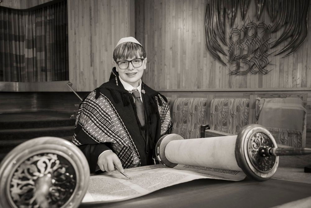 Young person in tallit and kippah reads from Torah scroll during Bar Mitzvah ceremony in synagogue.