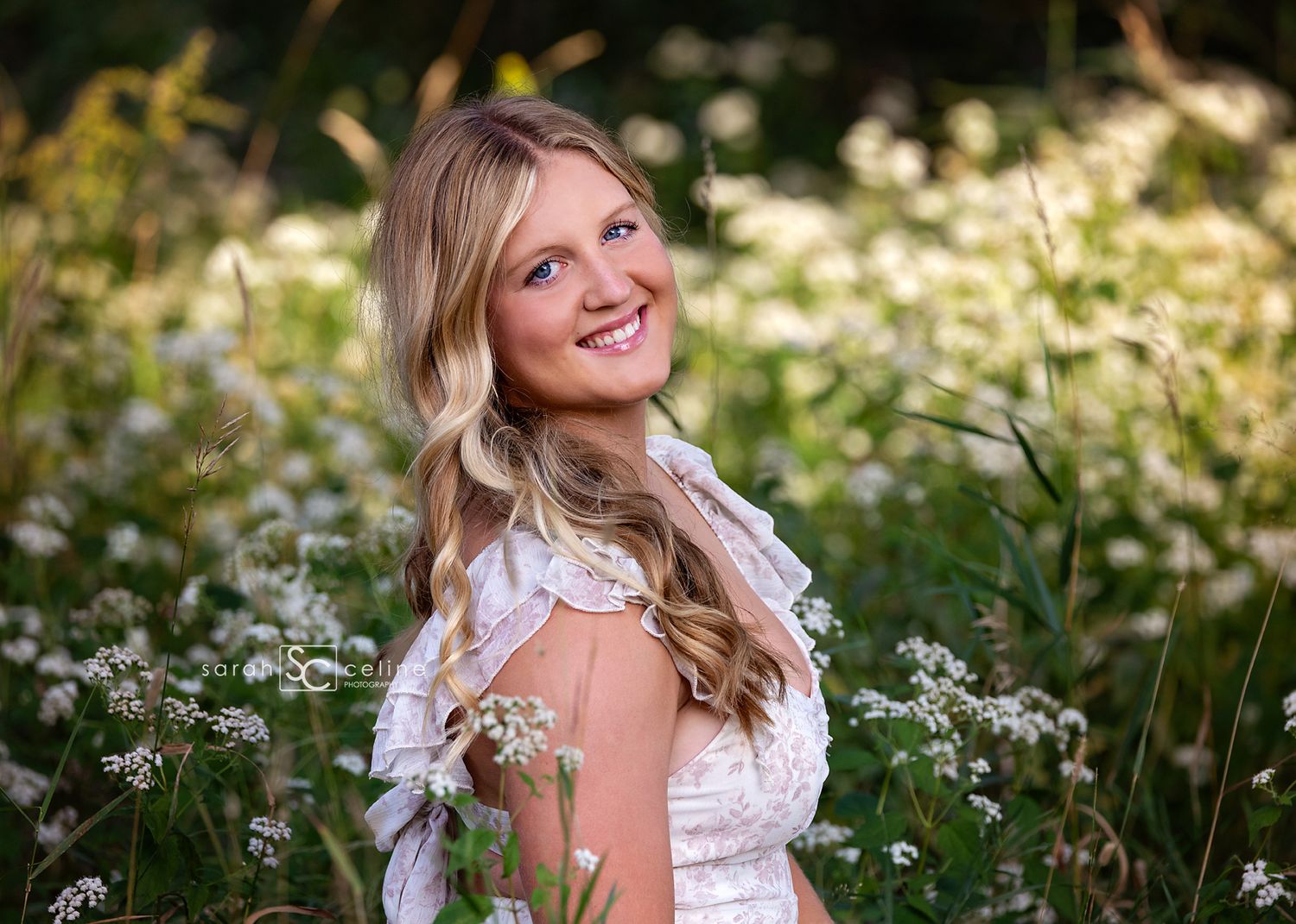 Smiling blonde woman in white lace dress posing among wildflowers in a sunlit meadow.