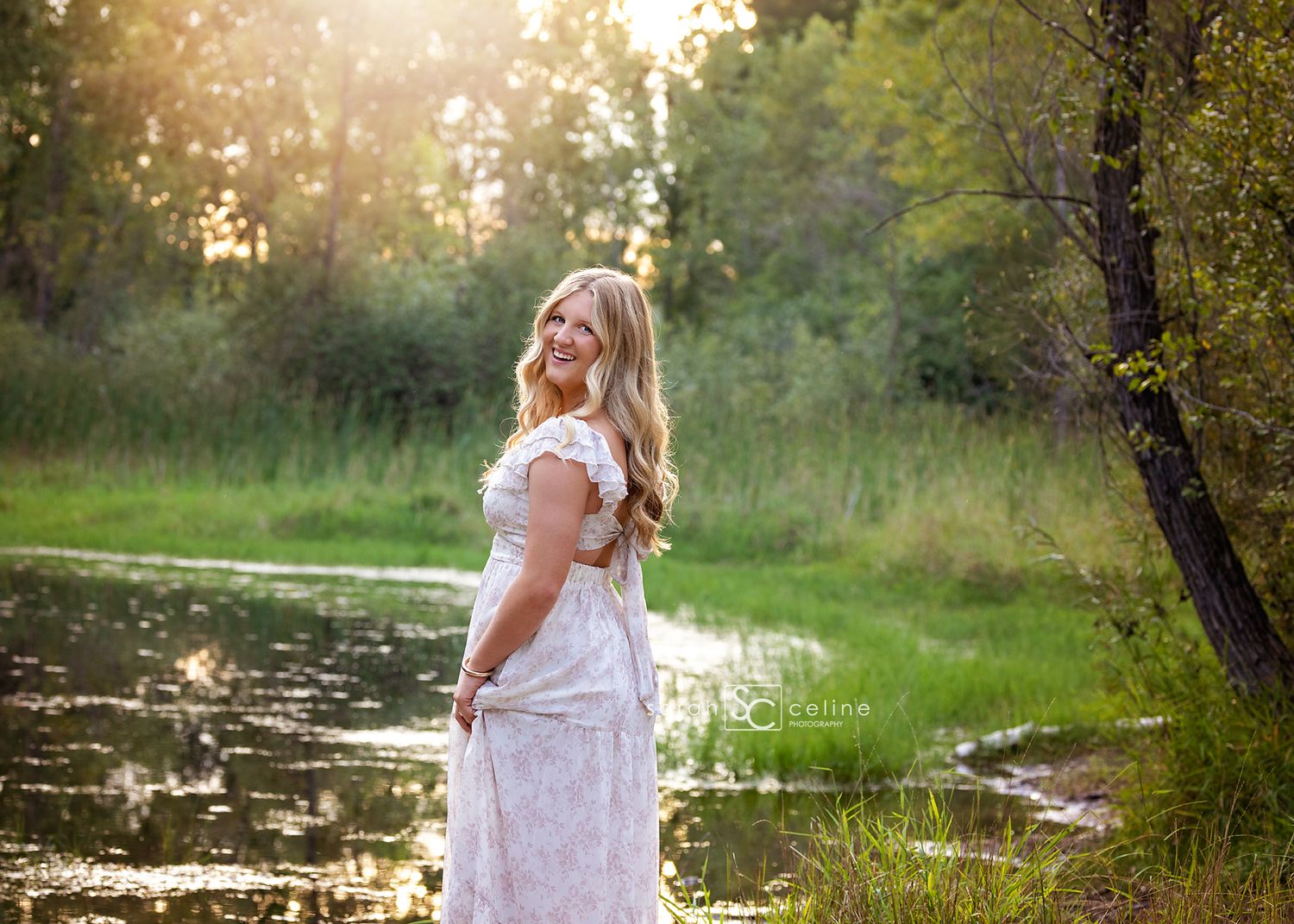 Pregnant woman in white lace dress posing by a serene pond surrounded by lush green trees in golden hour light.