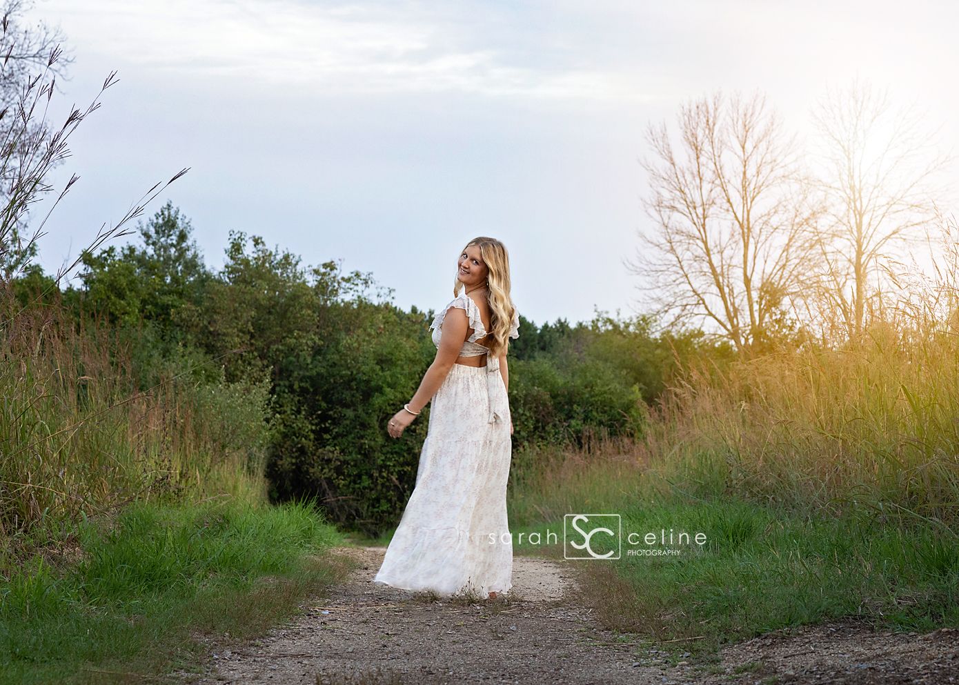 A bride in a flowing white lace gown walks down a rustic dirt path surrounded by lush greenery at golden hour.