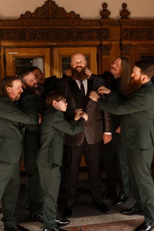 Groom in brown suit smiles as groomsmen in dark green suits help him get dressed before wedding ceremony.
