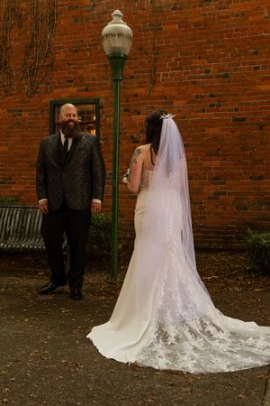 Bride in white lace gown and veil faces groom in dark patterned suit near brick wall and vintage lamp post.