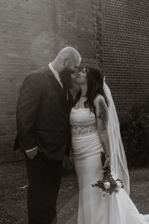 Black and white photo of a tattooed bride and groom sharing a kiss near a brick wall on their wedding day.