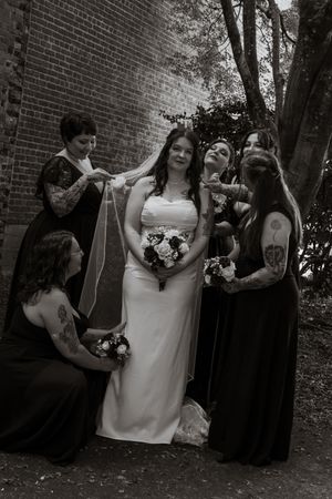 Black and white photo of a bride in white gown holding bouquet surrounded by bridesmaids in dark dresses near brick wall.