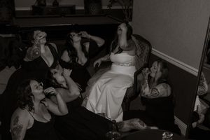 Black and white photo of a bride in white dress celebrating with bridesmaids in black dresses on a couch.