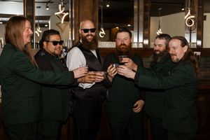 Six men in dark green suits raising glasses in a toast inside an elegant bar with warm lighting.