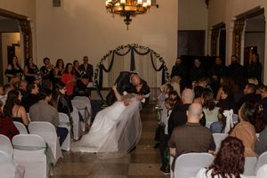 Bride and groom share a romantic dip kiss at indoor wedding ceremony with seated guests and elegant chandelier.