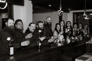 Black and white photo of a wedding party smiling and raising glasses at a bar in a celebratory toast.