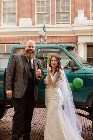 Bride and groom in wedding attire flipping off camera, standing before a teal truck on brick street.