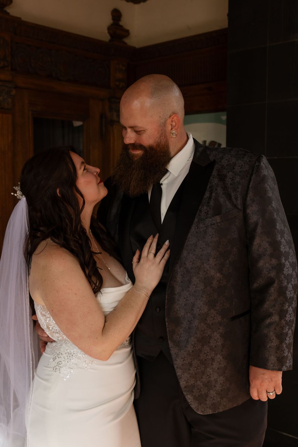 Bride and groom share an intimate moment, smiling at each other, groom in dark patterned tuxedo jacket.