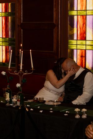 Bride and groom share a romantic kiss at candlelit wedding reception table with stained glass windows.
